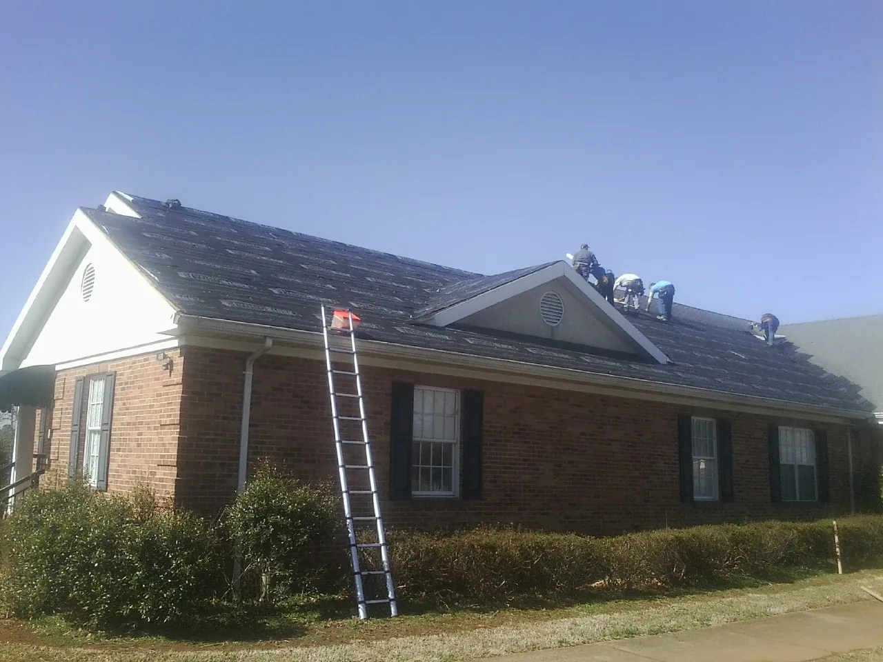 Crew tearing shingles off a brick ranch roof with a ladder set — Emergency Roof Repair work by our crew in Bailey