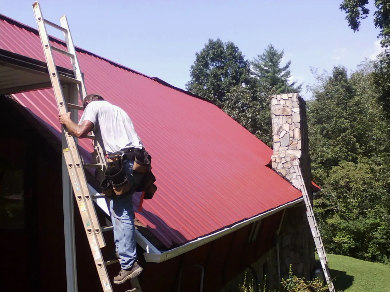 Roofer on a ladder beside a red metal roof and stone chimney — Soffit & Fascia Repair by Roofing Brothers in Bailey